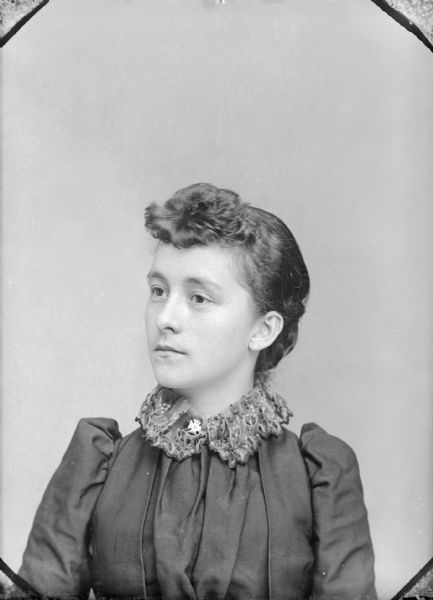 Quarter-length studio portrait of a woman. She is wearing a dark-colored blouse with a lace collar and a cross collar pin.