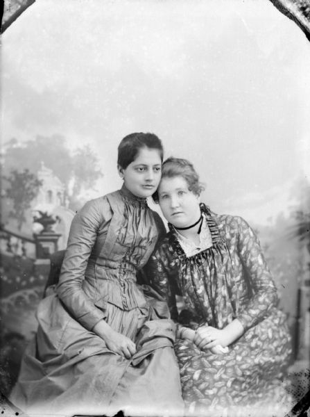 Studio portrait in front of a painted backdrop of two women posing sitting and leaning against each other. They are both wearing dark-colored dresses, with the woman on the right wearing a floral print dress and a ribbon around her neck.