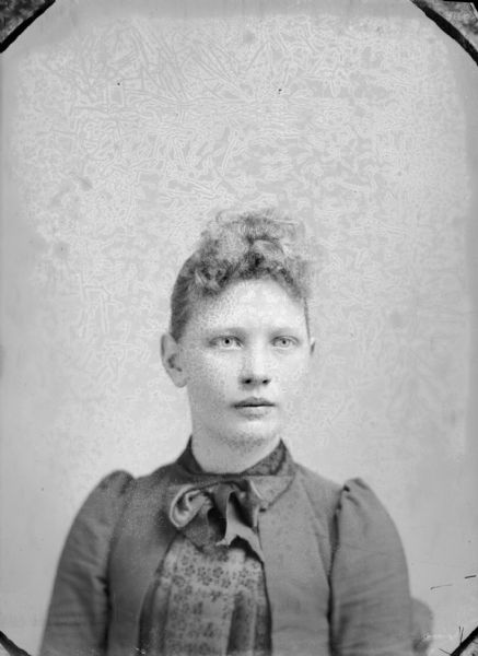 Quarter-length sudio portrait of a young woman posing sitting. She is wearing a dark-colored dress with a neck bow tie.