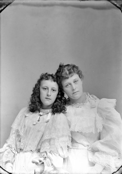 Studio portrait of two young women posing sitting. They are wearing light-colored dresses with lace trim. The younger woman on the left has shoulder-length curly hair.