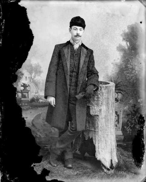 Studio portrait of man with a moustache posing standing and leaning with his left arm on a stump and holding a glove, and wearing a dark-colored overcoat, suit coat, vest, trousers, and a hat. The man is identified as P.H. Jones.