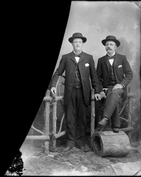 Studio portrait in front of a painted backdrop of two men posing with a prop fence and gate. The man with a moustache on the right is posing sitting with his legs crossed, and a man is posing standing on the left. Both men are wearing dark-colored suit coats, vests, bow ties, hats and trousers. The man on the right is probably Peter Moe.