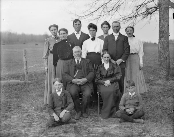 Outdoor group portrait of a family group, including five young women and two young men posing standing, an older man and woman are posing sitting in chairs, and two young boys are posing sitting on the ground. The group is identified as the family of George Thompson.