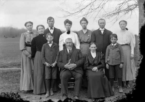 Outdoor group portrait of a family group, including five young women, two boys, and two young men posing standing. There is an older man and woman posing sitting in front in chairs. The group is identified as the family of George Thompson.