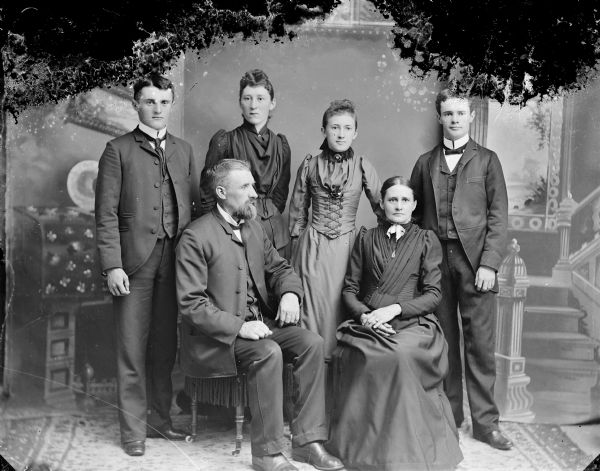 Studio group portrait of two young women and two young men posing standing behind an older man and woman sitting in chairs. The family group is identified as the family of George Hull.