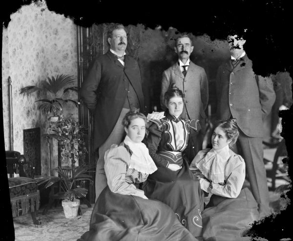 Studio group portrait of three men posing standing behind a woman posing sitting in a chair, flanked by twins posing sitting on the ground. The persons are identified as the following, from left to right, as: top row, unidentified, John Mills, unidentified/obscured by emulsion damage, bottom row, a Spaulding twin, Alice Jones — the wife of John Mills, and a Spaulding twin.