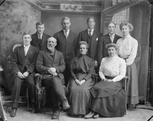 Studio portrait in front of a painted backdrop of a family group, including four men and a woman posing standing behind two men and two women posing sitting. Identified as the family of J.D. Perry, and individually as the following: back row, Charlie Perry, Harriet Perry, Austin Perry, Archie Perry, and Gem or Heon Perry. front row Howard Perry, J.D. Perry, the wife of J.D. Perry, and Ella Perry.