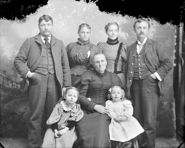 Studio portrait in front of a painted backdrop of two European American men and two European American women posing standing, and an European American woman posing sitting in the center with two European American girls. Identified as possibly the Maddocks and Roddy families.