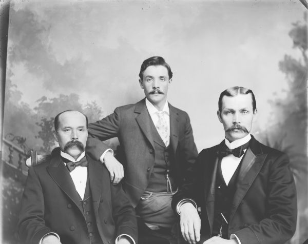 Studio group portrait in front of a painted backdrop of three European American men posing sitting. Identified as the Samdahl/Sanidahl men, probably brothers. Identified, left to right as: Simon Samdahl, Olaf Samdahl, and Ed Samdahl.
