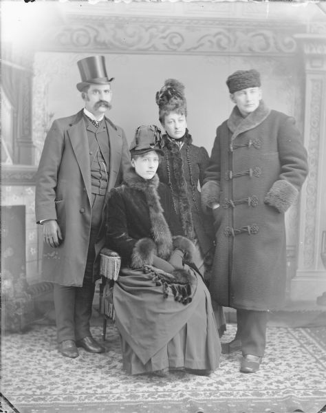 Studio group portrait in front of a painted backdrop of two European American men and two European American women. Identified, from left to right, as: Frank Long, Lottie Porter Long (sitting), Alice Porter Whitting, and Al Whitting.The men reportedly took over the Porter drugstore from their father-in-law, and it became the Whitting-Long drugstore.