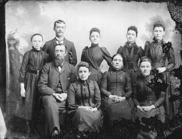 Studio group portrait in front of a painted backdrop of the Henry Steihl Family. The man with a beard posing sitting on the left iis dentified as Henry Steihl, and Ella Steihl and Joe Steihl are posing standing in the upper left.