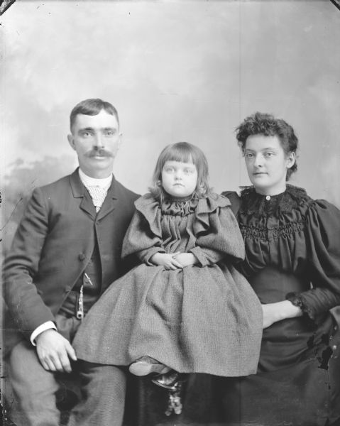 Studio group portrait in front of a painted backdrop of Mr. and Mrs. Philander Hill, and a girl sitting between them, probably their daughter.