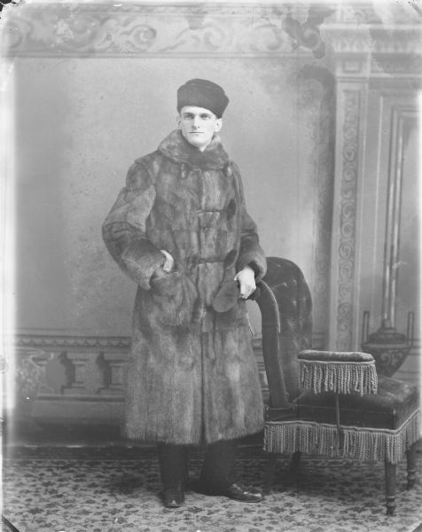 Full-length studio portrait in front of a painted backdrop of a man posing standing near an upholstered chair. He is wearing a long fur coat and hat.