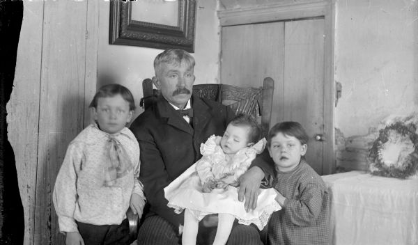 Interior of a room with a European-American man posing sitting and holding a deceased small European-American girl holding a flower in her lap. One either side of the man and girl are two European-American boys. On the right is a table covered by a light-colored cloth and a floral wreath. There is also a wooden door, and a framed picture on the wall.