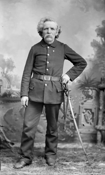 Full-length studio portrait in front of a painted backdrop of man with a moustache. He is standing and holding a sword with his left hand, and is wearing a belt over a button-down coat. The man is identified as Charles Schenk.