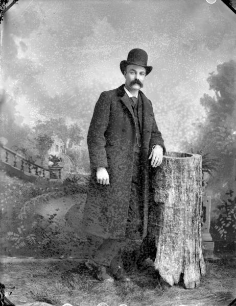 Full-length studio portrait in front of a painted backdrop of a European American man, with a moustache, posing standing with his legs crossed and leaning with his left arm on a tree stump. He is wearing a hat, long overcoat, and suit coat. Identified as Professor J.H. Persey/Dersey, who was reportedly in Black River Falls in 1897.