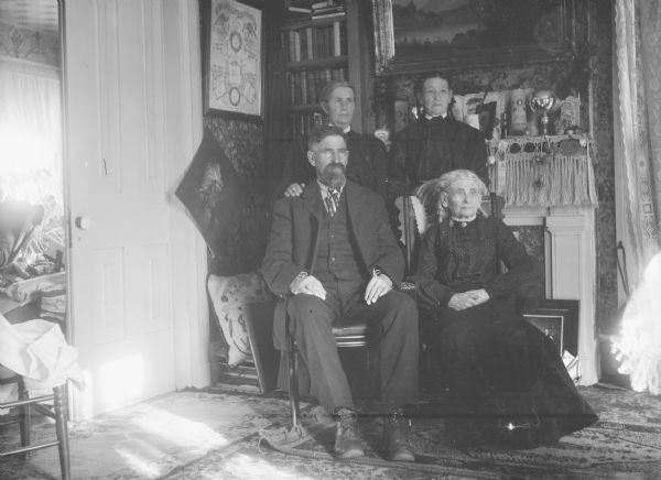 Indoor group portrait of a man and three women. The woman sitting on the right is identified as Mrs. Calvin Johnson. The items on the mantle and in room are used for identification of location.
