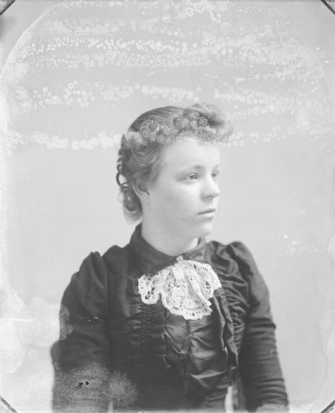 Waist-up studio portrait of a woman with blond hair posing sitting. She is wearing a dark-colored dress and a light-colored lace neck detail.