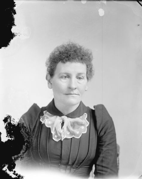 Studio portrait of a woman posing sitting. She is wearing a dark-colored dress and light-colored lace neck bow.