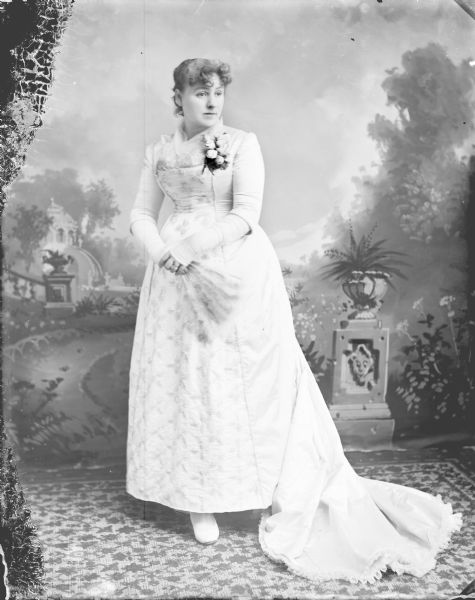 Full-length studio portrait in front of a painted backdrop of a woman posing standing. She is wearing a light-colored wedding dress and is holding a hand fan.