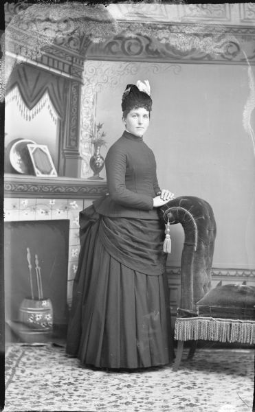 Full-length studio portrait in front of a painted backdrop of a European American woman posing standing. She has her hands on the back of a chair and is wearing a dark-colored dress and hat.
