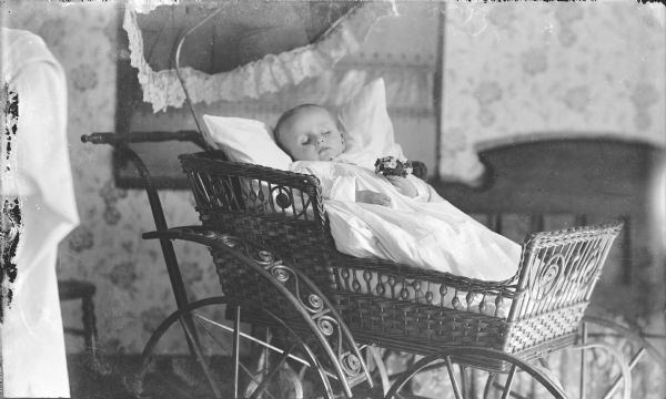 Indoor portrait of a post-mortem infant lying in a baby buggy and holding a bunch of flowers.