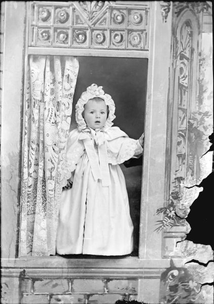 Studio portrait of a small girl wearing a dress and bonnet. She is posing standing in a prop window.