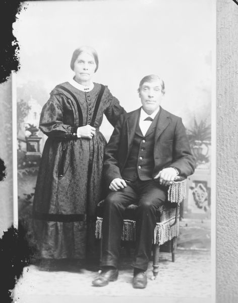 Copy photograph of a studio portrait of an European American woman posing standing on the left and an European American man posing sitting on the right.