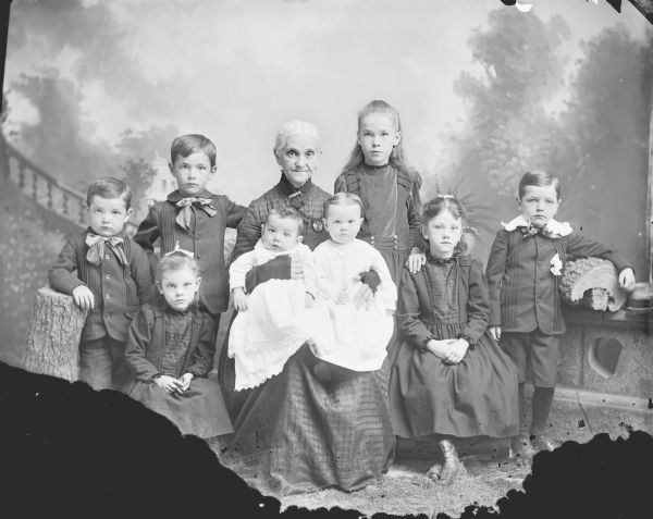 Studio group portrait in front of a painted backdrop. In the center, an elderly woman is sitting and holding two infants in her lap, and three girls, and three boys are standing around her.