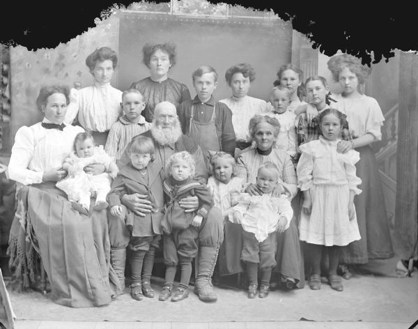 Studio group portrait in front of a painted backdrop of a family. The group includes eight women, one man, six boys, two girls, and an infant.