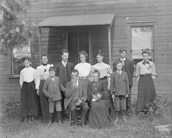 Outdoor group portrait of a European American family sitting and standing outdoors on the lawn in front of a house. The group includes three men, six women, and two boys.