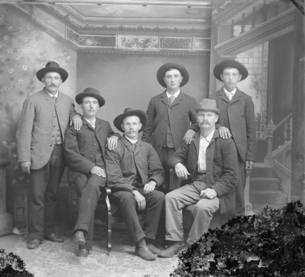Studio group portrait in front of a painted backdrop of six men posing sitting and standing. They are all wearing suit coats and hats.