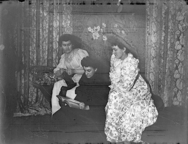 Interior group portrait of three European American woman posing sitting and looking at a book. The woman on the left is knitting.