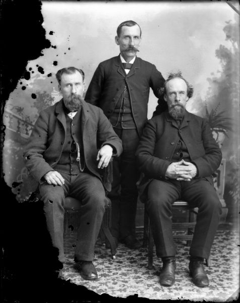 Studio group portrait in front of a painted backdrop of two men posing sitting in front of a man posing standing between them.