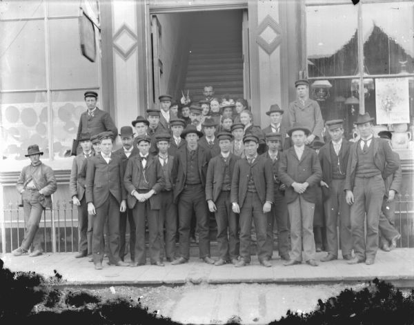 View from street of a group of twenty-eight men and seven women posing standing in front of a doorway between two store windows. Location identified as Main Street, between Jones Lumber and Merchantile and W.C. Jones grocery store, directly in front of the entrance to the office of Dudley Spaulding and Port's Hall. The individuals are identified in the front row, standing, left to right: 2nd. Oliver Darwin; 3rd. (a little behind) Jeff Peasley.
