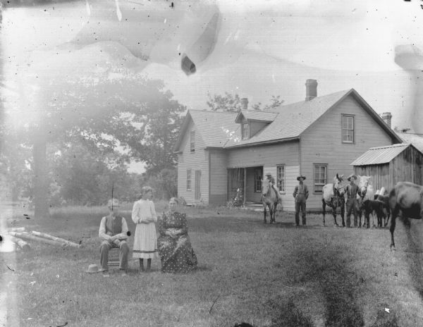 View across lawn in an unidentified location of a girl posing standing on the left between a man and woman posing sitting in the yard of a farmhouse. On the right are two young men, one displaying two horses, and a boy posing sitting on a horse.