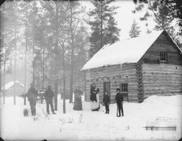 View towards two men standing on the left, two women in the center who are holding infants in their arms, and two boys standing on the right. The group is posing standing on the snow-covered ground in front of log cabins.