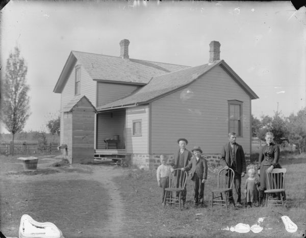 Outdoor group portrait in an unidentified location of a man, woman, three boys, and a girl posing standing behind three empty wooden chairs. Behind them is a two-story wooden house with a stone foundation, small porch, and a small structure that may be an outhouse.