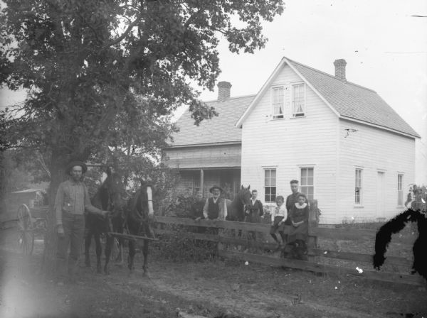Outdoor group portrait in an unidentified location of a group of men, women and children posing on the lawn and road in front of a two-story wooden house. On the far left is a man displaying two horses hitched to a wagon. Behind the fence is a man and woman standing. Another woman is standing behind the group in the center holding the bridle of a house. On the right is a woman standing behind a boy and girl sitting on the wooden fence.