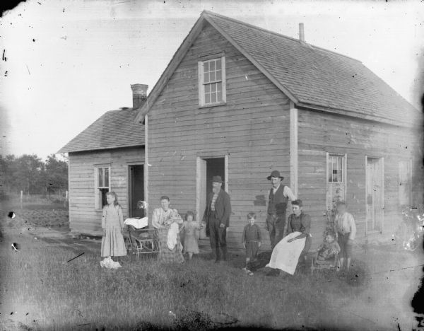 Exterior portrait in an unidentified location of a large group, including two men posing standing, two women posing sitting, one woman holding an infant, three two boys and two girls posing standing and sitting.