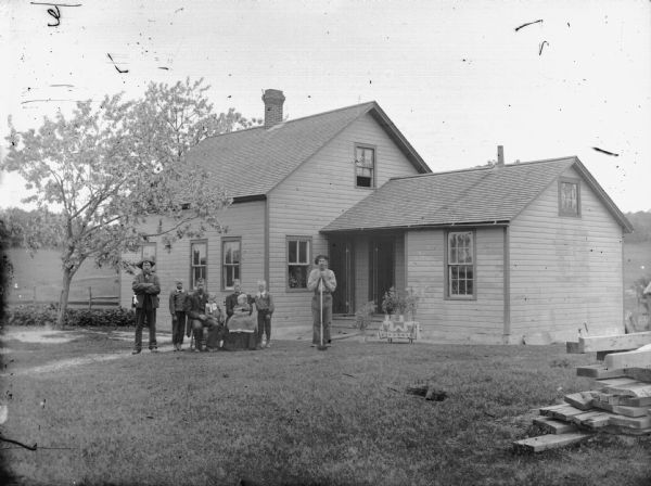 Outdoor group portrait in an unidentified location of a group posing in the yard of wooden house. There is a man and a woman sitting in the center, each holding a child in their lap. Standing on either side of them are two boys and two men. A pile of lumber in the foreground on the right.