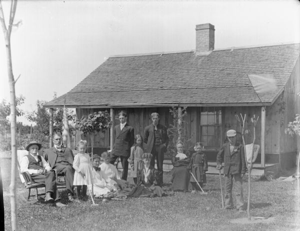 Outdoor group portrait in an identified location of a group of European American people posing in the yard of a small wooden cottage. There are two men and a woman sitting in chairs, three women are sitting on the ground, two men are standing, three girls are standing, and a boy is standing. Two of the girls and the one boy are holding croquet mallets. A flag is flying from the porch, and another is on a flagpole behind the group on the lawn.