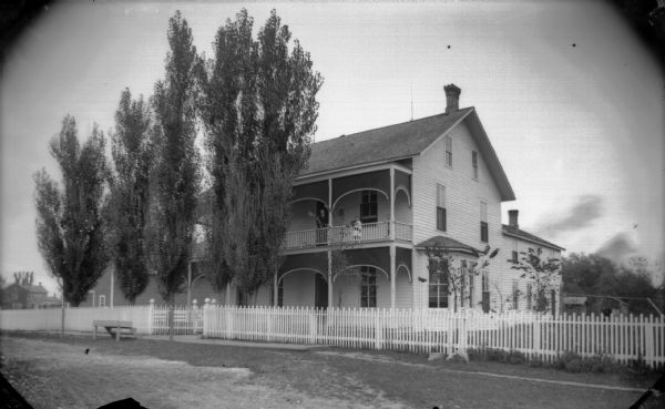 View across road or driveway towards a large white two-story wooden building with a porch and balcony, and a fence and gate bordering the yard. Wooden steps are near the gate facing the road. A European American woman and two children are posing on the second floor balcony. Identified as 326 South Second Street, and the residence of H.A. Bright, formerly the United States Hotel.