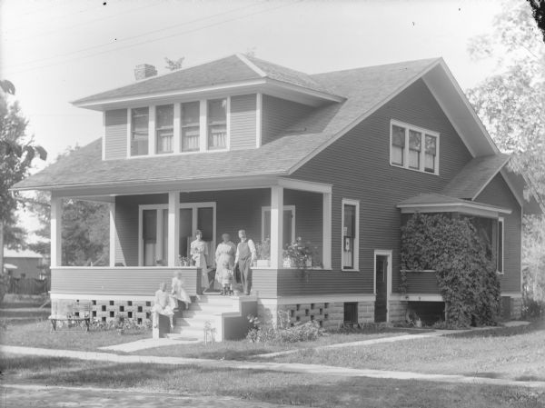 View across street towards a group of European Americans posing on the entry steps and porch of a two-story wooden house. A man, two women, and a child are standing on the porch, and two girls are sitting on the side of the steps. Identified as 118 South 5th Street, the home of Joe Carnahan.