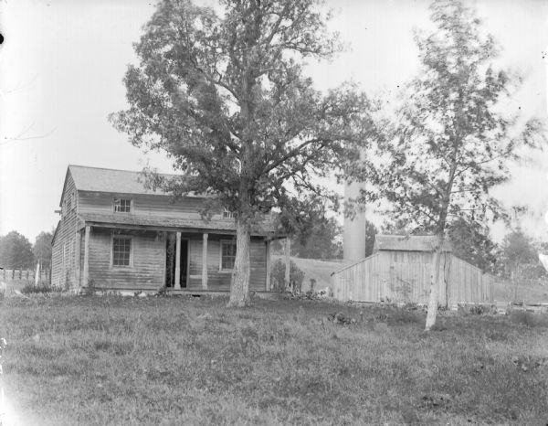 View across yard towards a two-story wooden house, with a wooden outbuilding on the right, with a tall water pipe behind it. Identified as the residence of John Coleman, near 10th Street and Monroe Street and Lake's Nursery.