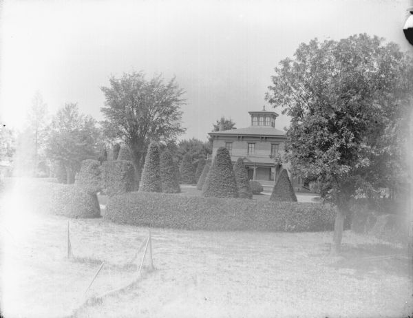 Slightly elevated view across lawn with a well-manicured yard with sculpted shrubs towards a three-story wooden house. Identified as the Spaulding residence.