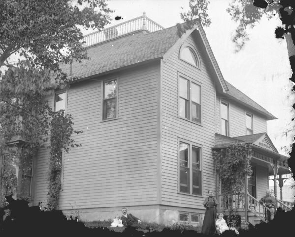 Outdoor group portrait of people in front of a large two-story wooden house, with a widow's walk on the roof. On the right a woman is standing on the steps leading up to the porch. On the lawn in front is a woman standing next to a girl sitting in a chair or infant carrier. In the center is a man reclining on the lawn with an infant. Location identified as 119 South Third Street, the residence of the Maddocks family.