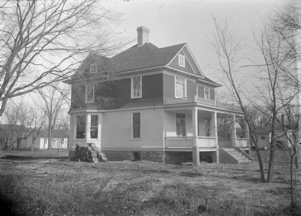 View across lawn towards the left side of a two-story wooden house, identified as 819 Main Street, the home of the Monsos Family. The front of the house has a porch and a balcony. A stack of wood is on the left side of the house. Buildings are in the background.
