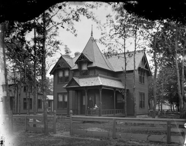 View across fence towards of a two-story wood-frame house with a porch. A man and an infant in a baby carriage are standing on the porch. The home is identified as the residence of Petra Kjorstad. There is another house on the left.