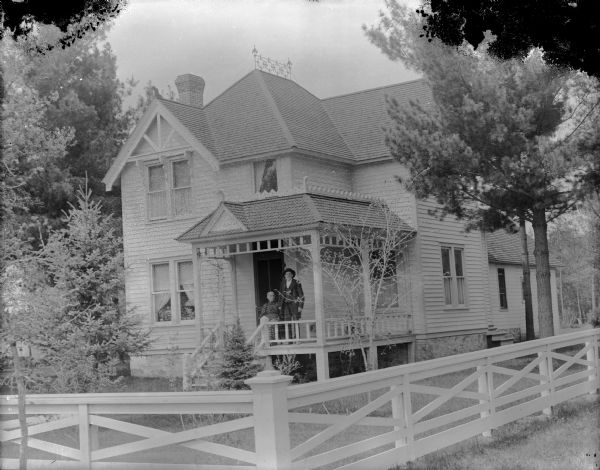 View across fence towards a man and woman posing on the porch of a two-story wood-frame house. The home is identified as the residence of Frank Cooper at 407 Monroe Street.